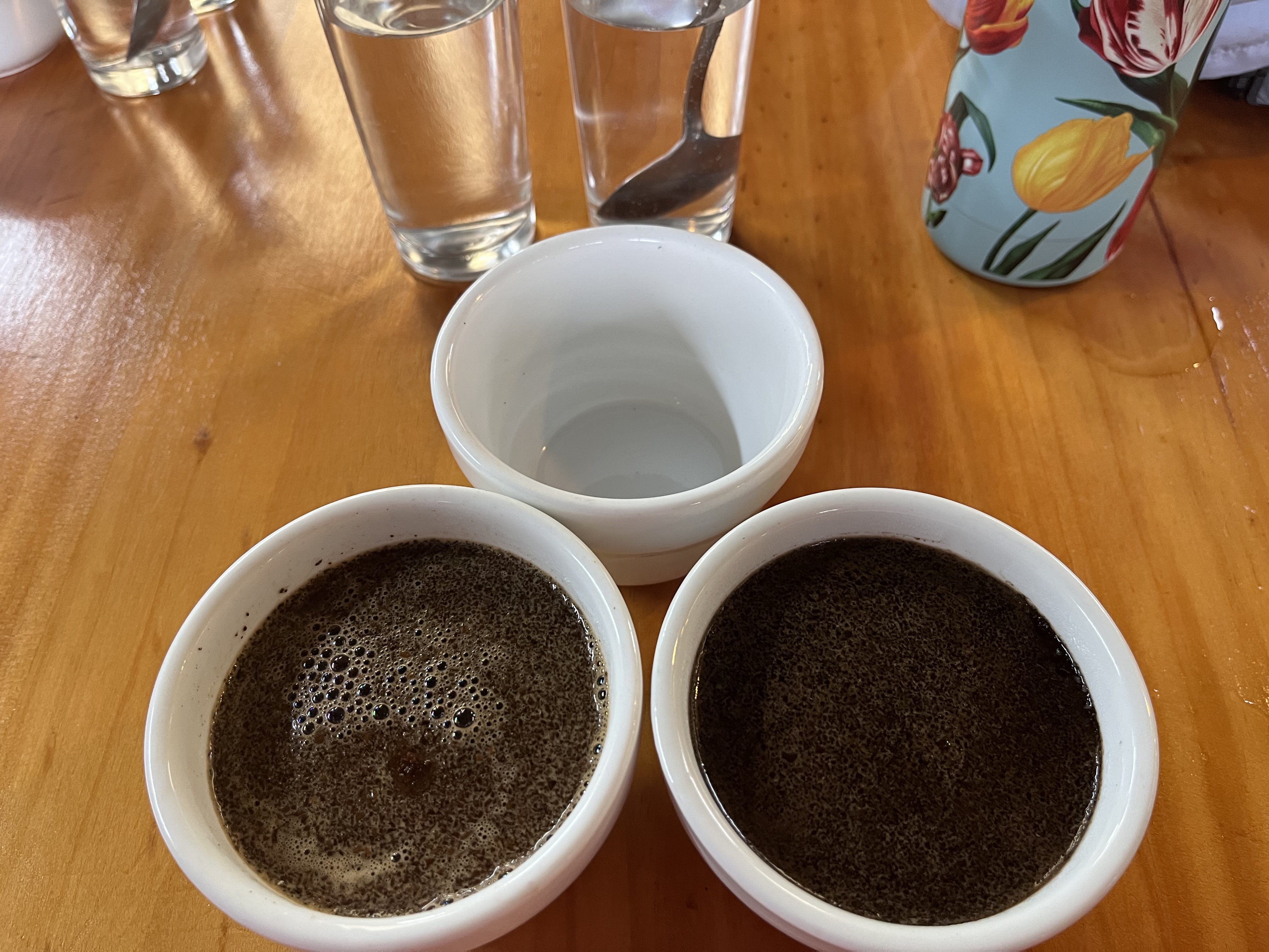 Two cups of brewed coffee prepared for tasting, showing light- and dark-roast samples on a wooden table with water glasses nearby.